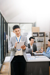 Young attractive Asian male office worker business suits smiling at camera with working notepad, tablet and laptop documents in office
