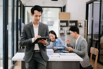 Young attractive Asian male office worker business suits smiling at camera with working notepad, tablet and laptop documents in office