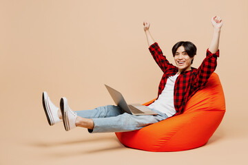 Full body young IT man of Asian ethnicity wear red shirt casual clothes sit in bag chair hold use work on laptop pc computer do winner gesture isolated on plain pastel light beige background studio.