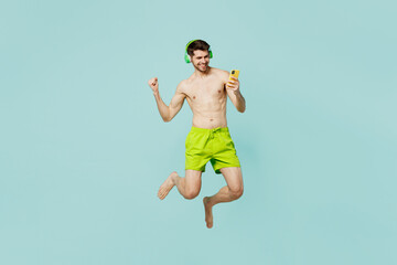 Fototapeta premium Full body side view young man in green shorts swimsuit headphones relax near hotel pool jump high use mobile cell phone listen music isolated on plain blue background. Summer sea rest sun tan concept