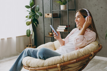 Obraz premium Young woman wears casual clothes sits in armchair listen to music in headphones use mobile cell phone stay at home hotel flat rest spend free spare time in living room indoor Lifestyle lounge concept