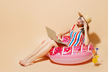 Full body young bored woman in one-piece swimsuit straw hat near hotel pool sit on donut ring use laptop pc computer isolated on plain light beige background Summer vacation sea rest sun tan concept