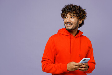 Young smiling Indian man wears red orange hoody casual clothes hold in hand use mobile cell phone...