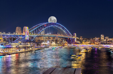 Obraz premium Panoramic night view of Sydney Harbour and City Skyline with a Sturgeon full Moon NSW Australia bright neon lights reflecting off the water