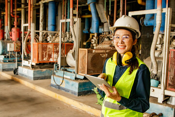 Asian female technician worker in engineering department wearing hard hat supervising machinery refrigeration system factory holding tablet checking control system standing and looking at work place.