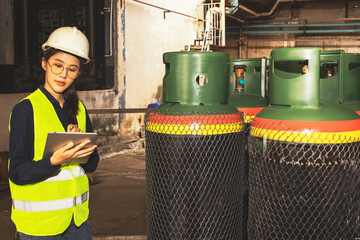 Asian female technician engineer inspector in industrial factory using digital tablet to inspect cylinders containing ammonia chemical refrigerant safe and well ventilated storage area for safety.