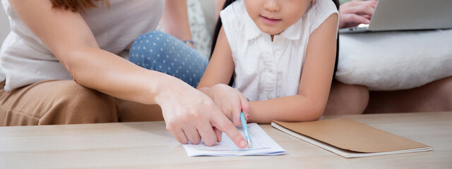 Happy family with mother teaching homework with daughter and father working with laptop on sofa in living room at home, mom explaining schoolwork with kid together, lifestyles and education.