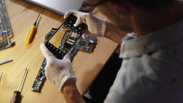 Novice computer repairman photographs details of motherboard at workplace with phone, over the shoulder. Student studies structure of laptop motherboard, photographs microchips.