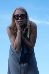 The woman is standing. The woman has a gray-brown dress, earrings, sunglasses. The woman has a blue scarf in her hand. The sky in the background.