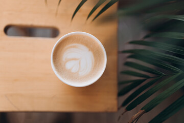Top view of a paper cup of coffee with latte art on wooden table. Street coffee, top view. Cup coffee in palm branches and leaves. Delicious hot beverage.