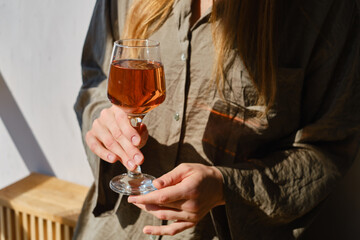 Glass of rose wine in the hands of a girl in the rays of the setting sun at home. Tasting of alcoholic beverages. Celebrate and enjoy the moment. Romantic evening aperitif. Close-up glass of wine
