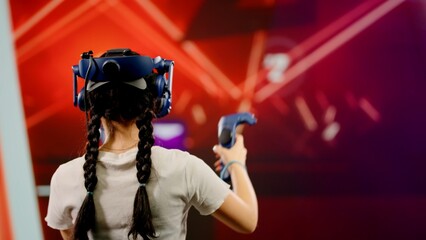 Obraz na płótnie Canvas The child use tracking controllers in her hands, moves actively immersing herself in modern game. The little girl wearing VR headset plays video game. Behind girl there is screen with blur background