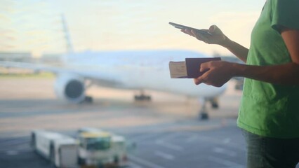 In an international airport, a young Caucasian woman stands by a large window, holding her passport and checking her flight details on her mobile phone. She's using an app and sending messages.