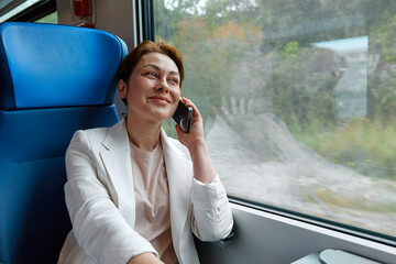 Smiling businesswoman freelance blogger using mobile phone in train. Pretty woman tourist traveling by train sitting near window and talking on smartphone.