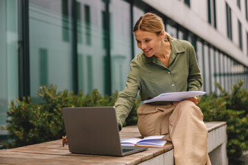 Woman analyst working with documents and use laptop sitting outside on modern building background