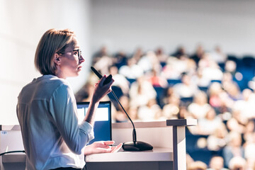 Female speaker giving a talk on corporate business conference. Unrecognizable people in audience at...