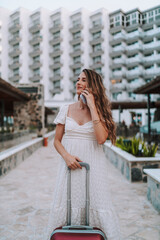 Chica joven delgada de vacaciones con maleta de viaje y smartphone paseando por la piscina de un hotel