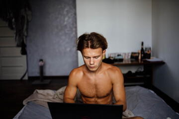 man with a naked torso works on the Internet with a laptop in bed at home.