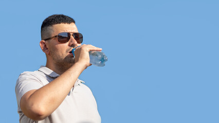 adult man drinking water from bottle in heat. guy suffering from thirst in against blue sky. prevention of dehydration on sunny day. thirsty male drink liquid for hydration, wellness, health. banner