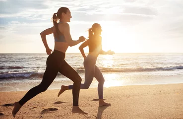 Tuinposter Cheerful active pretty young caucasian women enjoy morning cardio workout, jogging on sea beach, full length © Prostock-studio