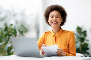 Happy black female student engaging in distance learning via laptop computer, holding copybook and smiling at camera