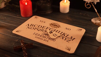 Close up shot of a wooden ouija board with a planchette on a table surrounded by candles, candlesticks and other elements in a muffed light.