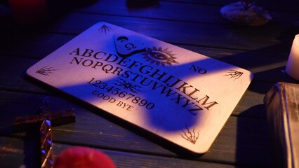 Close up shot of a wooden ouija board with a planchette on a table surrounded by candles, candlesticks and other elements in a muffed light. A shadow from a cross falls down on the board.