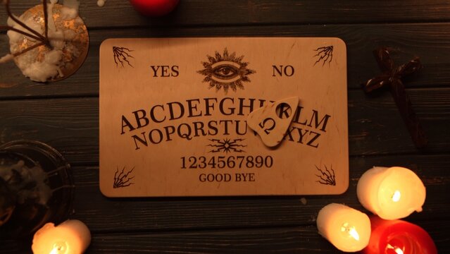 Close up shot of a wooden ouija board with a planchette on a table surrounded by candles, candlesticks and other elements in a muffed warm light.