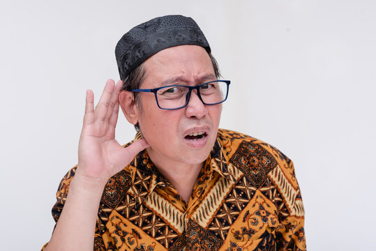 A deaf indonesian man asks someone to speak louder. Gesturing with hand on ear. Wearing a batik shirt and songkok skull cap. Isolated on a white background.