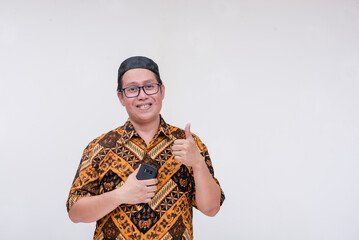 A middle aged man looking at the camera and making thumbs up sign while holding his cellphone. Wearing a batik shirt and songkok skull cap. Isolated on a white background.