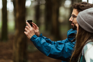 Cheerful young caucasian couple in jackets in forest enjoy travel vacation, make photo on smartphone