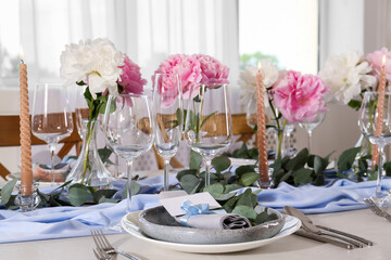 Beautiful table setting. Plate with greeting card, napkin and branch near glasses, peonies, burning candles and cutlery on table in room