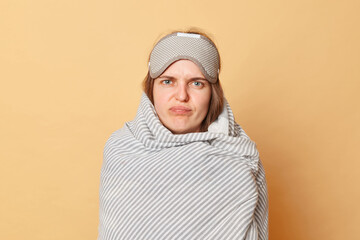 Sad unhappy upset woman wrapped blanket and wearing sleeping eye mask isolated over beige...