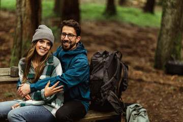 Cheerful young caucasian couple in jackets in forest, enjoy travel, vacation sit on bench with backpacks, hugging