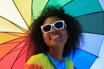 Portrait   LGBTQ  black woman holding a small heart  ball with sunglasses and a rainbow umbrella on a sunny day  at the bright tropical beach