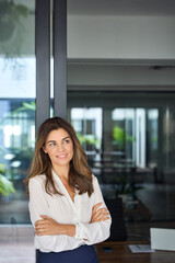 Fototapeta na wymiar Smiling dreamy Latin professional mid aged business woman corporate manager leader looking away, happy mature female executive standing at office window arms crossed, thinking. Vertical