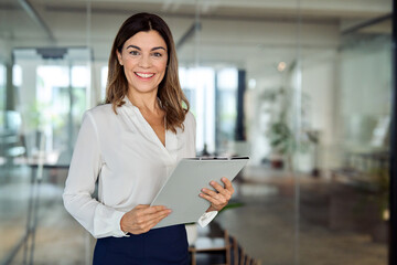 Happy 45 years old Latin professional middle aged business woman corporate leader, happy mature female executive, lady manager standing in office holding clipboard looking at camera, portrait.