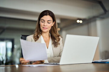 Busy 40 years old business woman working in office checking documents. Mid aged businesswoman accounting manager executive or lawyer using laptop reading paper file financial report, tax invoice. - obrazy, fototapety, plakaty