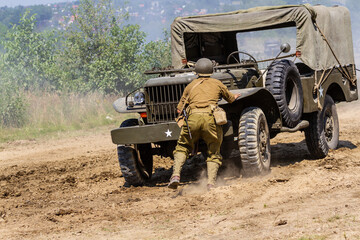 Fototapeta premium Historical reconstruction. An American infantry soldier from the World War II prepare to attack hidden behind a milirary vehicle between smoke and dust. View from the back.