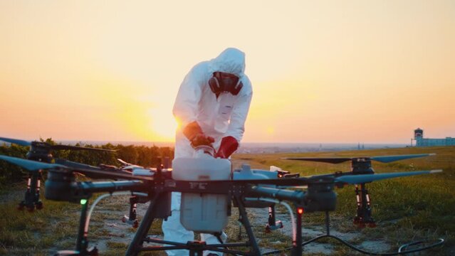 Male farmer in a protective white suit pours chemicals into the tank of an agricultural drone for spraying in a meadow at sunset