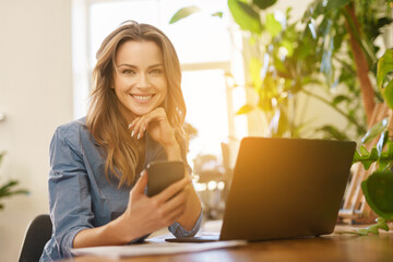 Beautiful smiling freelance woman in blue shirt working on laptop in a sunlit room filled with plants