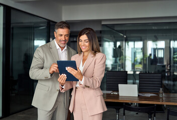 Two busy older professional corporate business executives man and woman wearing suits holding tablet technology device having discussion working on digital project plan standing in office at meeting. - 637057711