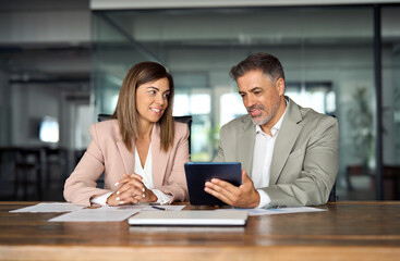 Two professional business executives working on tablet at office meeting. Managers team using digital pad computer sitting at table. Mature colleagues company board discussing smart tech solutions.