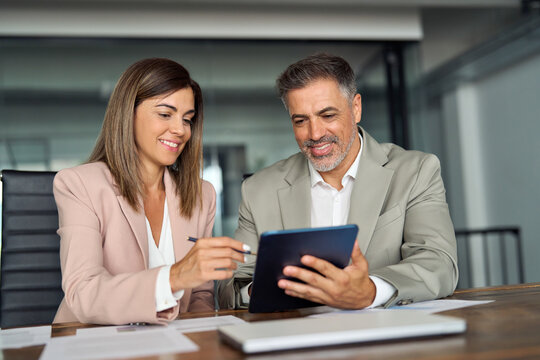 Two professional middle aged executives business team people talking working in corporate office at meeting having conversation looking at digital tablet company device discussing sales project.