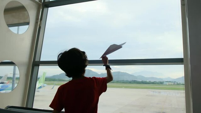 Children stand in front of a large airport window, playing with a paper airplane, their imaginations soaring as they pretend to be pilots. Young travelers, creativity, and the excitement of summer 