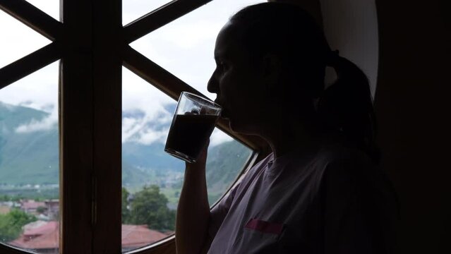 Close-up of silhouette of young woman looking out the window while drinking coffee, enjoying amazing view of mountain village outside, slow motion