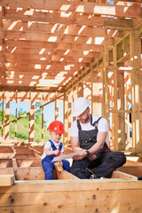 Fototapeta na wymiar Father with toddler son constructing wooden frame house. Male builder showing his son the construction plan, wearing helmets and blue overalls on sunny day. Carpentry and family concept.
