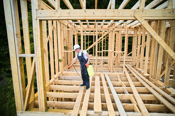 Laborer constructing wooden frame house near forest. Man treating woods, applying fire retardant using sprayer, while dressed in protective suit, helmet. Concept of modern eco-friendly construction.