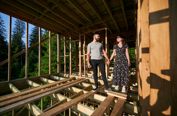 Fototapeta premium Man and woman inspecting their future wooden frame dwelling nestled near the forest. Youthful couple at construction site in early morning. Concept of contemporary ecological construction.