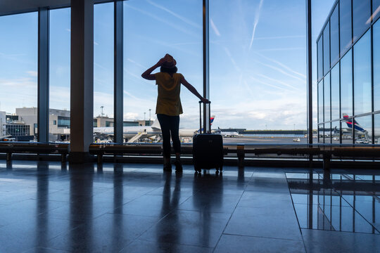 Woman passenger with luggage of a suitcase in the airport terminal looking at the planes from the window